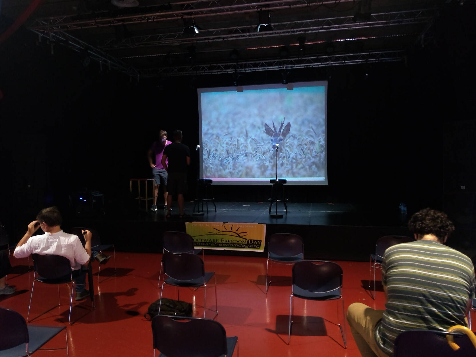 Sala amb cadires i un escenari amb una pantalla gran. A la pantalla hi ha la foto d'un cabirol. Dos ponents es preparen per fer una xerrada.